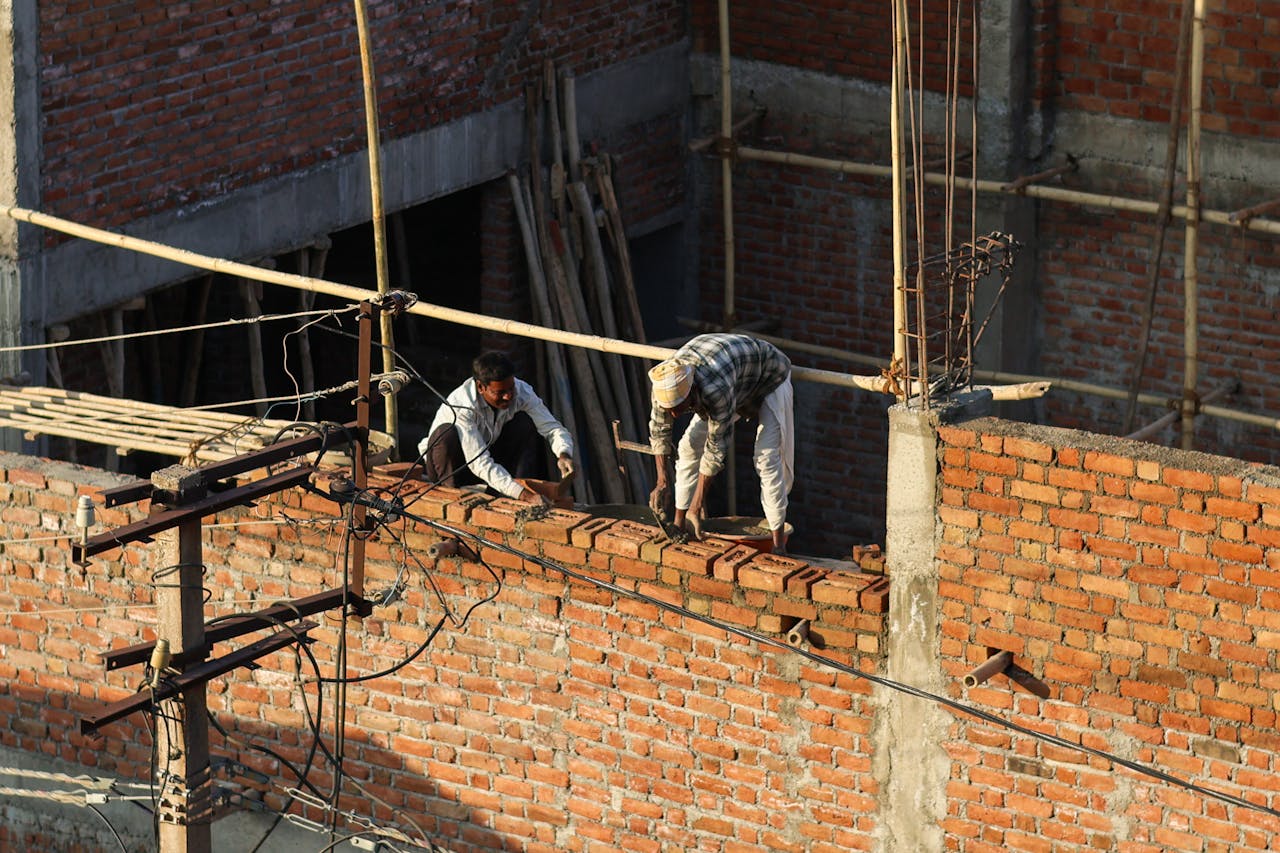 Two workers building a brick wall on a construction site under sunlight.